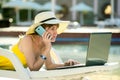 Young woman on beach chair at swimming pool working on computer laptop and talking on sell phone in summer resort. Remote work and Royalty Free Stock Photo