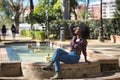 Young woman, beautiful and black with afro hair, with flower shirt, jeans and boots, sitting in a fountain playing with her hair. Royalty Free Stock Photo