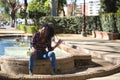 Young woman, beautiful and black with afro hair, with flower shirt, jeans and boots, sitting in a fountain playing with her hair. Royalty Free Stock Photo
