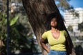 Young woman, beautiful and black with afro hair, with yellow shirt and jeans, leaning on the trunk of a tree in a sensual and