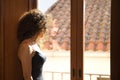 Young woman, beautiful, brunette, with curly hair, in a black dress, looking absentmindedly out the window of a room in a Royalty Free Stock Photo