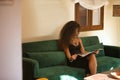 Young woman, beautiful, brunette, with curly hair, in a black dress, sitting on the sofa in a hotel lounge, reading a magazine. Royalty Free Stock Photo