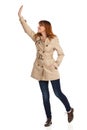 Young Woman In Beige Coat, Jeans And Brown Shoes Is Looking Away And Waving Hand Royalty Free Stock Photo