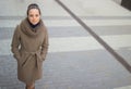 A young woman in a beige coat walks thoughtfully down the street