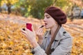 Young woman in beret wearing lipstick and looks at phone. Autumn Park background. Beauty girl