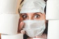 Young woman with big scared eyes in two medical virus protection face masks looks through stacks of toilet paper. Covid-19 Royalty Free Stock Photo