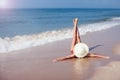 A young woman in bikini and in straw hat lying on a tropical beach, stretching up slender legs. Blue sea in the background. Royalty Free Stock Photo