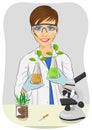 Young woman biologist in white coat holding two flasks with plants next to microscope in laboratory