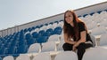 A young woman in black clothes with long hair is sitting on a stadium bleachers alone and and watching a sport game under the Royalty Free Stock Photo