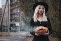 Young woman in the black coat holding the halloween pumpkin with the white smoke coming from inside of it in the autumn