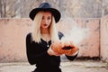 Young woman in the black coat holding the halloween pumpkin with the white smoke coming from inside of it in the autumn