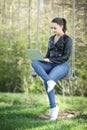 Young woman in a black jacked with coffee cup and laptop working outside in a park. Remote work. Digital work.