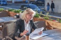 Young woman in black jacket and glasses sitting at a table in street cafe, looking through documents. Business activity, Royalty Free Stock Photo