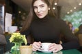 Young woman in black jersey look to camera, drink coffee and having rest in cafe near window. Royalty Free Stock Photo