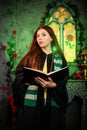 A young woman in a black mantle with a striped scarf around her neck casts a spell from a book and conjures with a wand of magic.