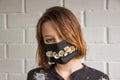 Young woman in the black medical mask with chamomile against the background of white bricks. Creative medical mask with flowers
