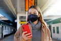 Young woman with black medical mask KN95 FFP2 using telephone in the train station with timetables on the background