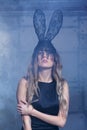 Young blonde woman in black rabbit or hare fancy mask and black dress. Female on smoke and metal wall background Royalty Free Stock Photo