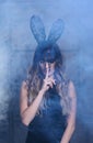 Young blonde woman in black rabbit or hare fancy mask and black dress. Finger on lips. Female on smoke and metal wall background Royalty Free Stock Photo