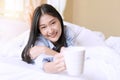 Young woman with blanket holding white cup of hot coffee or tea while lying on the bed in the morning after wake up Royalty Free Stock Photo