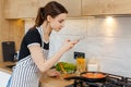 Young woman blogger in apron taking photo with phone while cooking food at kitchen. Preparing meal with frying pan on Royalty Free Stock Photo