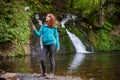 Young woman blogger in autumn mountains near lake and waterfall is recording video, taking photo with selfie stick Royalty Free Stock Photo