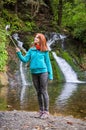 Young woman blogger in autumn mountains near lake and waterfall is recording video, taking photo with selfie stick Royalty Free Stock Photo