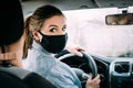 A young woman with blond hair in a black medical mask and a turquoise jacket is driving a left-hand drive