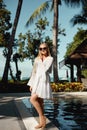 Young woman with blond hair in peignoir and bikini is posing around outdoor swimming pool in hotel resort with palm tree Royalty Free Stock Photo