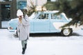 Young woman blonde girl in warm clothes with cup of coffee in hands near retro car decorated Christmas tree in winter street Royalty Free Stock Photo