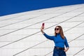 Young woman in blue clothes taking selfie photo by smartphone on background of huge concrete slabs of modern building.