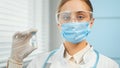 Young woman in blue disposable mask glasses and white coat holds transparent vial with vaccine