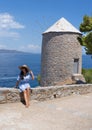 A young woman in a blue dress and a large straw hat is sitting next to old windmill in Greece