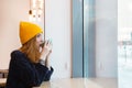 Young woman with blue eyes and blond hair in a yellow hat is drinking coffee in a cafe and looking into the window Royalty Free Stock Photo