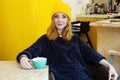 Young woman with blue eyes and blond hair in a yellow hat is drinking tea in a cafe near to yellow fridge Royalty Free Stock Photo