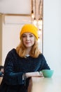 Young woman with blue eyes and blond hair in a yellow hat is drinking coffee in a cafe and looking into the camera. Royalty Free Stock Photo