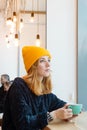 Young woman with blue eyes and blond hair in a yellow knitting hat is drinking coffee in a cafe. Royalty Free Stock Photo