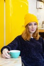 Young woman with blue eyes and blond hair in a yellow knitting hat is drinking coffee in a cafe. Royalty Free Stock Photo
