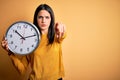 Young woman with blue eyes doing countdown holding big clock over yellow background pointing with finger to the camera and to you, Royalty Free Stock Photo