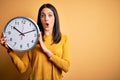 Young woman with blue eyes doing countdown holding big clock over yellow background scared in shock with a surprise face, afraid Royalty Free Stock Photo