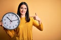 Young woman with blue eyes doing countdown holding big clock over yellow background with surprise face pointing finger to himself Royalty Free Stock Photo