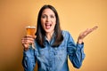 Young woman with blue eyes drinking glass of beer standing over isolated yellow background very happy and excited, winner Royalty Free Stock Photo