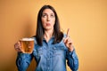 Young woman with blue eyes drinking jar of beer standing over isolated yellow background Pointing up looking sad and upset,