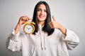 Young woman with blue eyes holding alarm clock standing over isolated white background happy with big smile doing ok sign, thumb Royalty Free Stock Photo