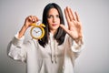 Young woman with blue eyes holding alarm clock standing over isolated white background with open hand doing stop sign with serious Royalty Free Stock Photo