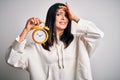 Young woman with blue eyes holding alarm clock standing over isolated white background stressed with hand on head, shocked with Royalty Free Stock Photo