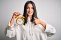 Young woman with blue eyes holding alarm clock standing over isolated white background with surprise face pointing finger to Royalty Free Stock Photo