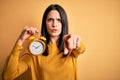 Young woman with blue eyes holding alarm clock standing over isolated yellow background pointing with finger to the camera and to Royalty Free Stock Photo