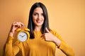 Young woman with blue eyes holding alarm clock standing over isolated yellow background with surprise face pointing finger to Royalty Free Stock Photo