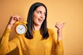 Young woman with blue eyes holding alarm clock standing over  yellow background pointing and showing with thumb up to the Royalty Free Stock Photo